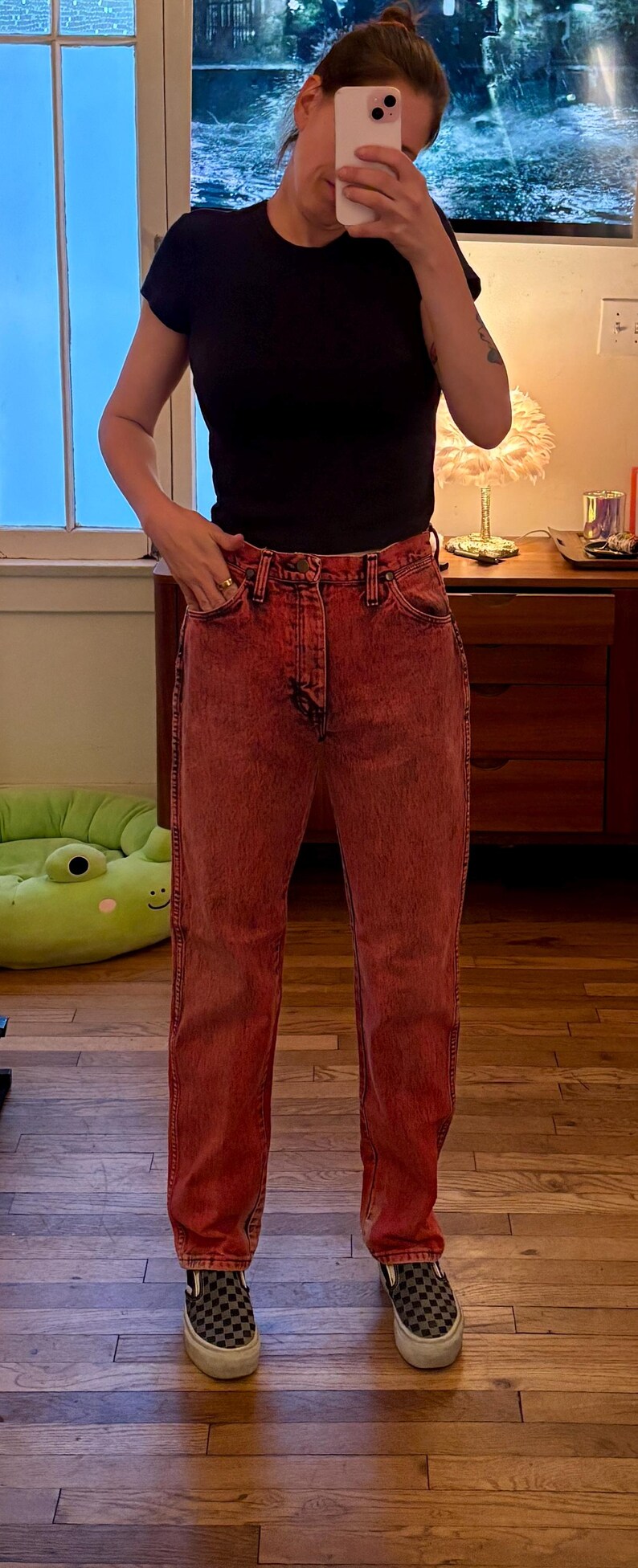 May include: A person wearing a black t-shirt and red acid wash jeans. The person is also wearing black and gray checkered slip-on shoes. The background includes a window and a wooden floor.