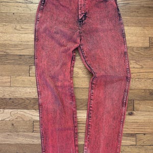 May include: Acid wash red denim jeans. The jeans have a high waist and a straight leg. The denim has a faded, textured appearance with darker red accents. The jeans are displayed on a wooden surface.