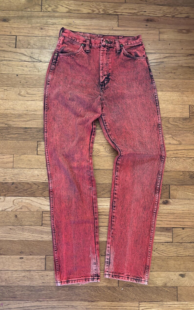 May include: Acid wash red denim jeans. The jeans have a high waist and a straight leg. The denim has a faded, textured appearance with darker red accents. The jeans are displayed on a wooden surface.