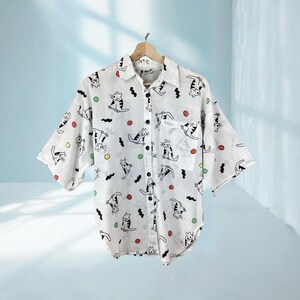 K&ouml;nnte beinhalten: Wei&szlig;es Button-Down-Hemd mit einem verspielten Katzenmuster. Das Hemd hat kurze &Auml;rmel, eine Brusttasche und einen Kragen. Die Katzenillustrationen sind schwarz, mit farbenfrohen Akzenten in Rot, Gr&uuml;n und Gelb. Das Hemd h&auml;ngt an einem Holzb&uuml;gel.