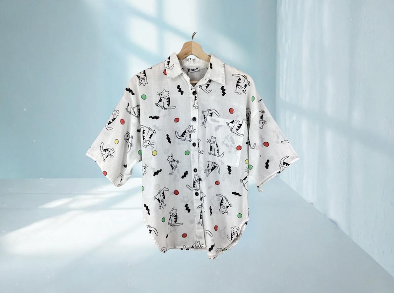 K&ouml;nnte beinhalten: Wei&szlig;es Button-Down-Hemd mit einem verspielten Katzenmuster. Das Hemd hat kurze &Auml;rmel, eine Brusttasche und einen Kragen. Die Katzenillustrationen sind schwarz, mit farbenfrohen Akzenten in Rot, Gr&uuml;n und Gelb. Das Hemd h&auml;ngt an einem Holzb&uuml;gel.