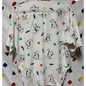 K&ouml;nnte beinhalten: Wei&szlig;es Hemd mit Kn&ouml;pfen und einem sich wiederholenden Muster aus Cartoon-Katzen in schwarzen Umrissen mit gestreiften Details. Das Design umfasst farbige Kreise und schwarze Zickzacklinien. Das Hemd hat kurze &Auml;rmel und einen Kragen.