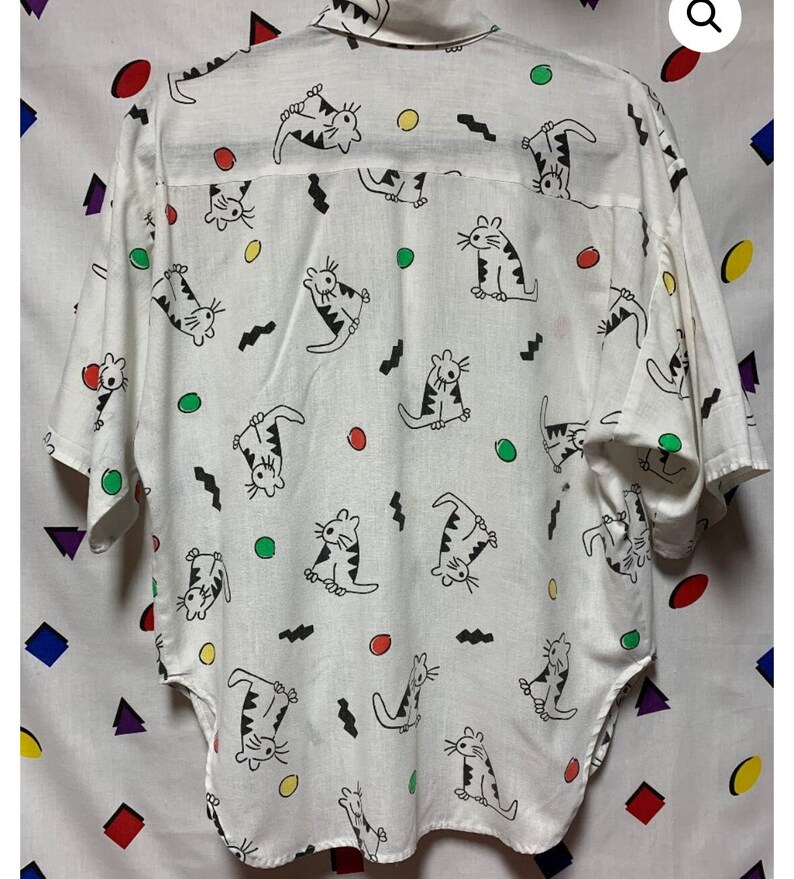 K&ouml;nnte beinhalten: Wei&szlig;es Hemd mit Kn&ouml;pfen und einem sich wiederholenden Muster aus Cartoon-Katzen in schwarzen Umrissen mit gestreiften Details. Das Design umfasst farbige Kreise und schwarze Zickzacklinien. Das Hemd hat kurze &Auml;rmel und einen Kragen.