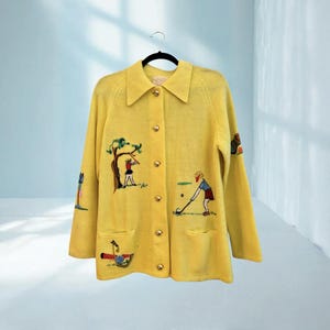 May include: A yellow cardigan with gold buttons and embroidered golf-themed designs. The cardigan features a collared neckline, two front pockets, and illustrations of golfers, trees, and golf equipment. The garment is hanging on a black hanger.