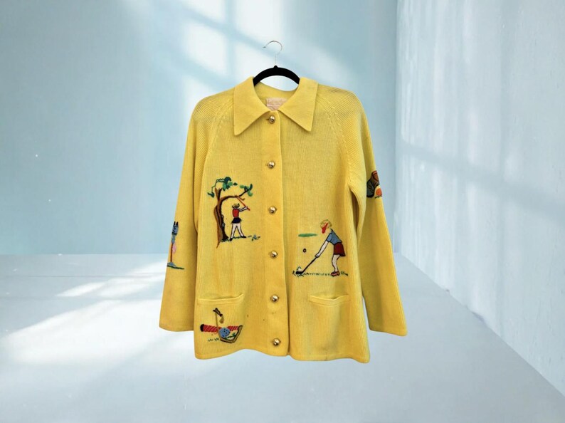 May include: A yellow cardigan with gold buttons and embroidered golf-themed designs. The cardigan features a collared neckline, two front pockets, and illustrations of golfers, trees, and golf equipment. The garment is hanging on a black hanger.