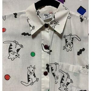 K&ouml;nnte beinhalten: Wei&szlig;es Hemd mit Kn&ouml;pfen und einem verspielten Katzenmuster in Schwarz. Das Hemd hat einen Kragen, eine Tasche und dunkelbraune Kn&ouml;pfe. Der Druck umfasst Cartoon-Katzen, schwarze Zickzacklinien und bunte Punkte. Das Etikett lautet "Cape Cod California".