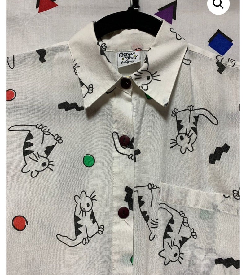 K&ouml;nnte beinhalten: Wei&szlig;es Hemd mit Kn&ouml;pfen und einem verspielten Katzenmuster in Schwarz. Das Hemd hat einen Kragen, eine Tasche und dunkelbraune Kn&ouml;pfe. Der Druck umfasst Cartoon-Katzen, schwarze Zickzacklinien und bunte Punkte. Das Etikett lautet "Cape Cod California".