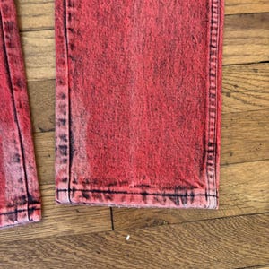 May include: Close-up of red acid wash denim jeans. The jeans have a faded, distressed look with black accents along the seams and edges. The fabric appears to be a medium weight denim.