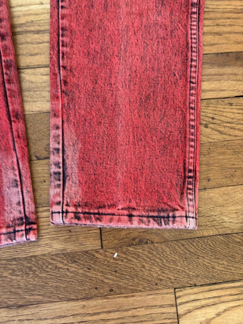 May include: Close-up of red acid wash denim jeans. The jeans have a faded, distressed look with black accents along the seams and edges. The fabric appears to be a medium weight denim.