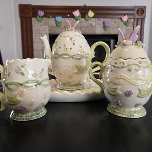 May include: A white ceramic mug with a black handle and the text "But First, Coffee" in a bold, black font. The mug is set against a blurred background, suggesting a cozy setting.