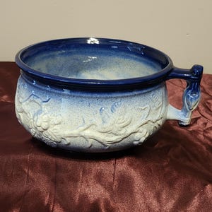May include: A large, blue and white ceramic pot with a single handle. The pot has a white glaze with a blue rim and a floral design.