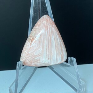 May include: A pale pink and white cabochon gemstone with a triangular shape. The gemstone is set in a clear plastic display stand.