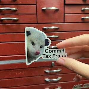 Puede incluir: Una pegatina de un opossum blanco y negro con el texto "Commit Tax Fraud" sobre un fondo transparente.