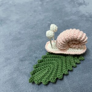 May include: A pink crocheted snail with white crocheted antennae sitting on a green crocheted leaf.