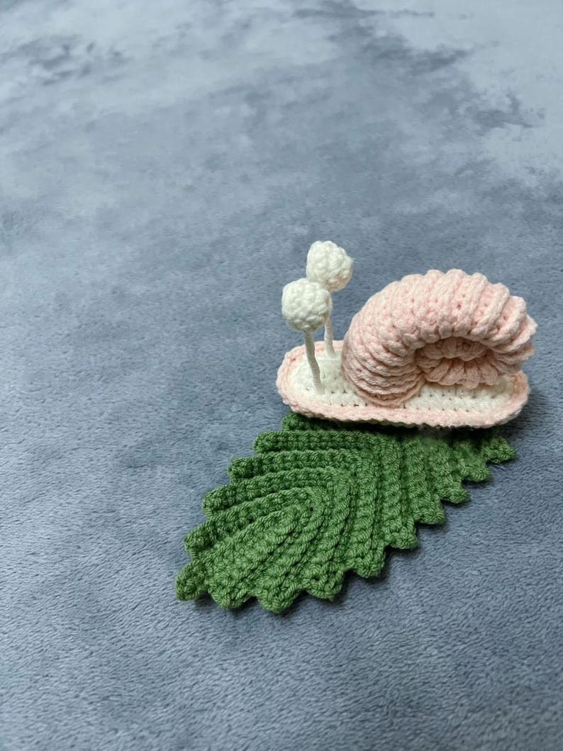 May include: A pink crocheted snail with white crocheted antennae sitting on a green crocheted leaf.