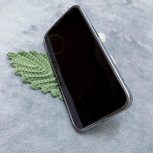 May include: A green crocheted leaf phone stand holding a black smartphone in a vertical position.