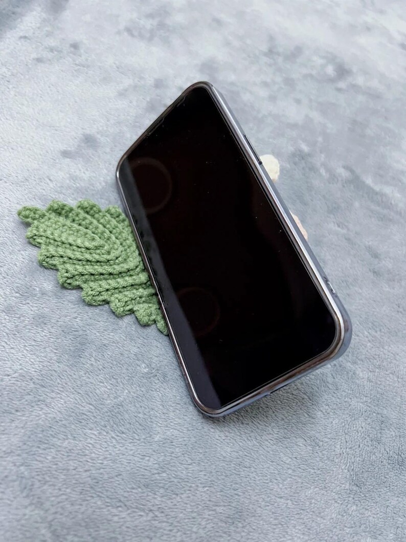 May include: A green crocheted leaf phone stand holding a black smartphone in a vertical position.
