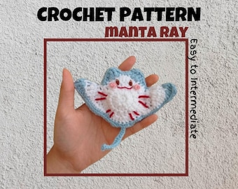 May include: A crocheted manta ray amigurumi toy with a blue and white body and red details. The toy has a smiling face with pink cheeks and is held in a hand. The image includes the text "CROCHET PATTERN MANTA RAY Easy to Intermediate".