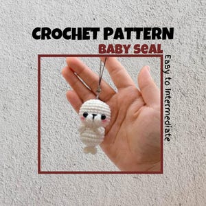 Puede incluir: Patrón de crochet para un amigurumi de foca bebé blanco. La foca está sosteniendo sus aletas frente a su cara y tiene ojos negros. El patrón se describe como fácil a intermedio.
