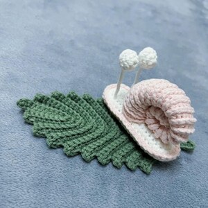 May include: A crocheted snail with a pink shell and white eyes, sitting on a green crocheted leaf.