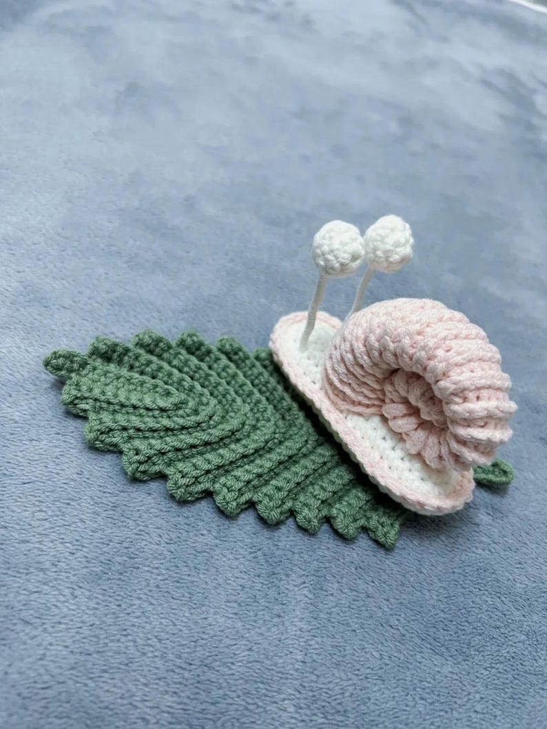 May include: A crocheted snail with a pink shell and white eyes, sitting on a green crocheted leaf.