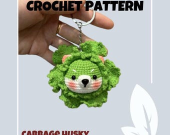 May include: Crochet pattern for a green cabbage husky keychain. The keychain is made of green yarn and has a white face with black eyes and a pink nose. The keychain is attached to a silver key ring.