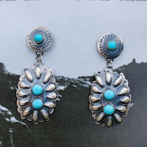 Könnte beinhalten: Ein Paar silberfarbene Ohrringe mit türkisfarbenen Akzenten. Jeder Ohrring hat eine kreisförmige Oberseite mit einem zentralen Türkisstein und ein hängendes, stilisiertes Blumendesign mit drei Türkissteinen. Die Ohrringe sind vor einem strukturierten Hintergrund.