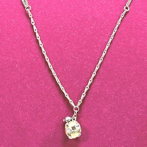 Puede incluir: Collar plateado con un pequeño colgante redondo. El colgante tiene un centro de color claro con un diseño decorativo y un pequeño lazo. El collar se muestra sobre una superficie rosa.