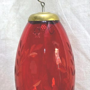 May include: A red glass Christmas ornament with a gold-colored metal cap and a silver hook. The ornament has an elongated teardrop shape and is decorated with etched leaf patterns. The ornament is hanging against a white background.
