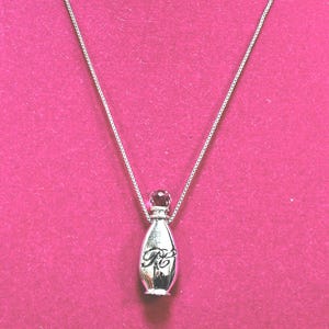 May include: A silver-colored perfume bottle pendant necklace. The pendant has a rounded shape with a small, dark-colored gem at the top and a decorative design on the front. The necklace chain is a delicate silver color.