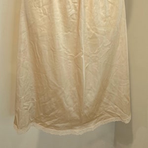 May include: A cream-colored half slip with a gathered waistband and a delicate lace trim at the hem. The slip is made of a smooth, silky fabric, and it hangs to about knee length.