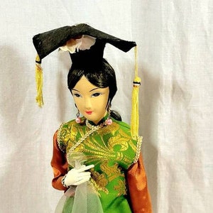 Könnte beinhalten: Eine traditionelle chinesische Puppe, die ein grünes und goldenes Brokat-Oberteil, einen durchsichtigen orangenen Rock mit goldenen Verzierungen und einen schwarzen Hut mit gelben Quasten trägt. Die Puppe steht auf einem schwarzen Sockel.