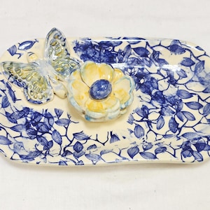 May include: A rectangular ceramic tray with a cream-colored base and blue floral patterns. A butterfly and a flower with yellow petals and a blue center are attached to the tray. The tray is approximately 8 inches long and 5 inches wide.