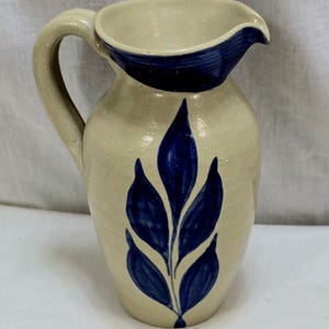 May include: A ceramic pitcher with a cream-colored base and a dark blue leaf design. The rim and handle are cream-colored. The pitcher has a spout for easy pouring and is approximately 8 inches tall.