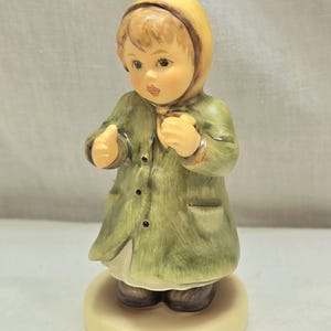 May include: A porcelain figurine of a young girl wearing a green coat, yellow hood, and brown shoes. She is holding her hands together in front of her.