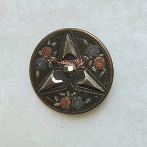 Puede incluir: Un botón redondo de latón con fondo negro y un diseño de estrella de tres puntas de plata. La estrella tiene un centro plateado elevado y facetado. El botón está decorado con flores pintadas en tonos rojos, naranjas, azules y verdes.