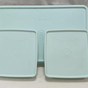 Op de afbeelding: Drie lichtblauwe Tupperware-deksels in verschillende maten. Eén groot rechthoekig deksel is achter twee kleinere vierkante deksels geplaatst. Elk deksel heeft een verhoogd Tupperware-logo en een gecentreerd cirkelvormig designelement.