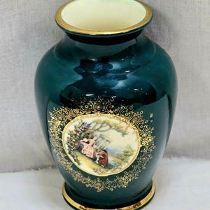 May include: A dark teal ceramic vase with a gold rim and base. The vase features a central oval design with a pastoral scene and gold filigree. The interior rim is cream-colored.