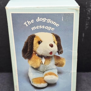 Könnte beinhalten: Ein Stoffhund-Spielzeug in einer Box. Der Hund hat braunes und beiges Fell, ein weißes Gesicht und trägt eine blaue Latzhose. Auf der Box steht der Text "The dog-gone message that I send..." auf blauem Hintergrund.