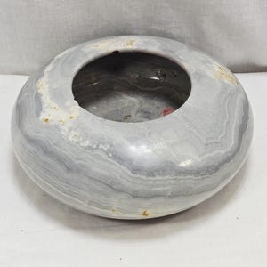 May include: A circular stone bowl in shades of grey and white, featuring a wide central aperture. The bowl's surface is polished, revealing layered patterns. The interior is dark, with subtle red and other colour accents.