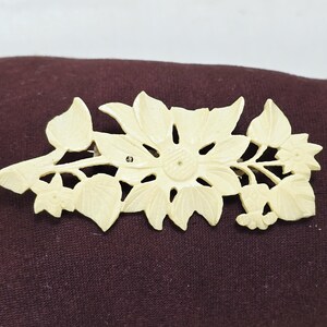 Peut inclure: Broche florale ivoire représentant un tournesol détaillé avec des feuilles et des tiges. La broche est faite d'un matériau de couleur claire et est posée sur un fond marron foncé. Un accessoire décoratif.