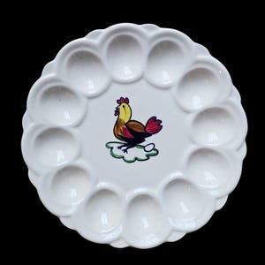 May include: White ceramic egg plate with a scalloped edge and 12 egg indentations. The center features a colorful illustration of a rooster on a green cloud. The rooster has yellow, brown, and red feathers.