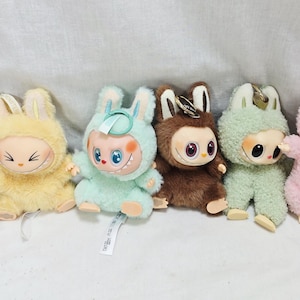 May include: A collection of six plush dolls, each featuring a unique color scheme and design. The dolls have large eyes, smiling faces, and bunny ear hoods. Colors include brown, yellow, mint green, and pink. Each doll has a keychain attached.