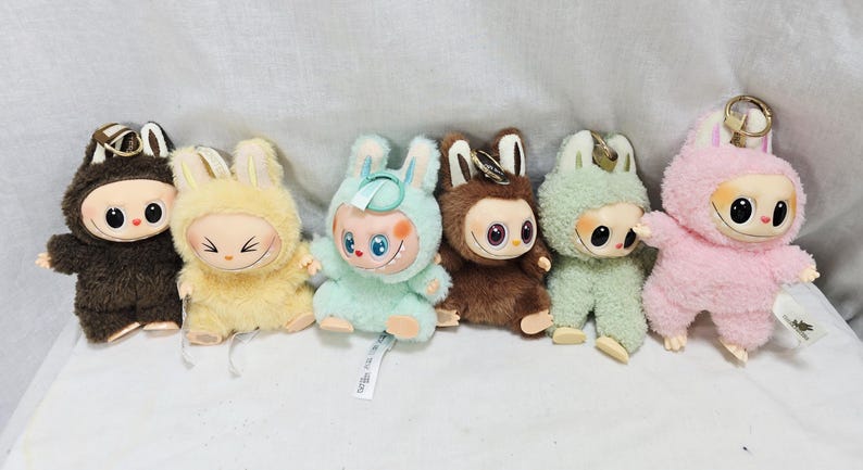 May include: A collection of six plush dolls, each featuring a unique color scheme and design. The dolls have large eyes, smiling faces, and bunny ear hoods. Colors include brown, yellow, mint green, and pink. Each doll has a keychain attached.