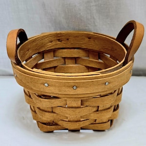 May include: A small, woven basket with a round shape and two leather handles. The basket is made of light brown wood and features a woven design. Small silver rivets secure the wooden bands. The handles are a lighter shade of brown.