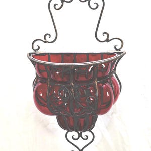 May include: A hanging planter with a deep red glass bowl encased in a black wrought iron frame. The frame features decorative scrollwork and a heart-shaped base. A purple ring is at the top for hanging. The planter is designed for indoor or outdoor use.