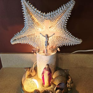 Pode incluir: Uma lâmpada decorativa com uma grande estrela-do-mar branca texturizada posicionada atrás de uma cruz central iluminada com um crucifixo prateado. Uma pequena estatueta com manto rosa e branco assenta numa base circular adornada com conchas.