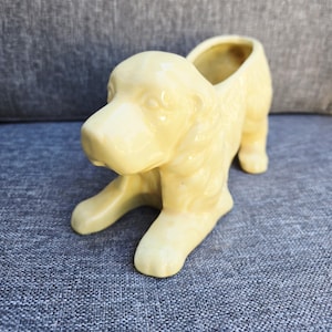 May include: A pale yellow ceramic dog figurine planter with a glossy finish. The dog is depicted in a lying position with its head raised and ears draped forward. The back of the dog features a hollow opening for small plants or items.