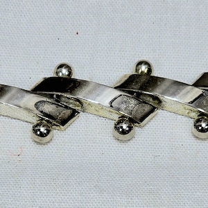 Puede incluir: Un brazalete plateado con un diseño en zigzag. Cada zig está conectado por una pequeña bola plateada redonda. El brazalete está hecho de un material metálico brillante y se muestra sobre una superficie blanca.