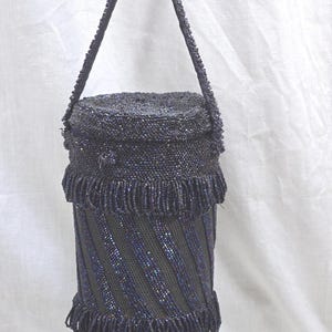 May include: A cylindrical, beaded handbag in dark blue. The bag features a beaded handle and is adorned with fringe at the top and bottom. The body of the bag has a diagonal striped pattern. The bag is likely vintage or handmade.
